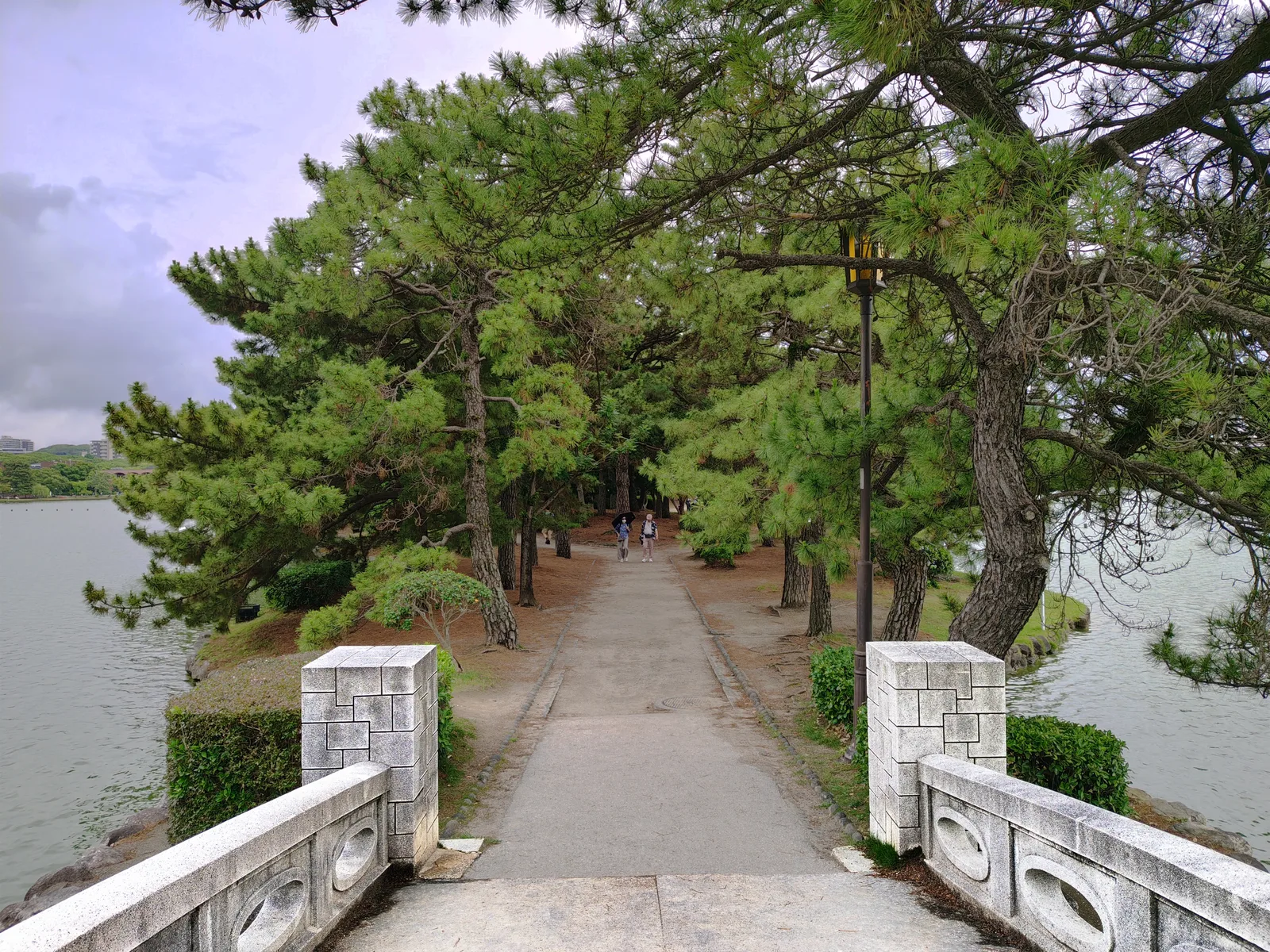 A stone bridge leading onto a pine-covered island in Ohori Park, with two people walking along the path and the park's lake on both sides.