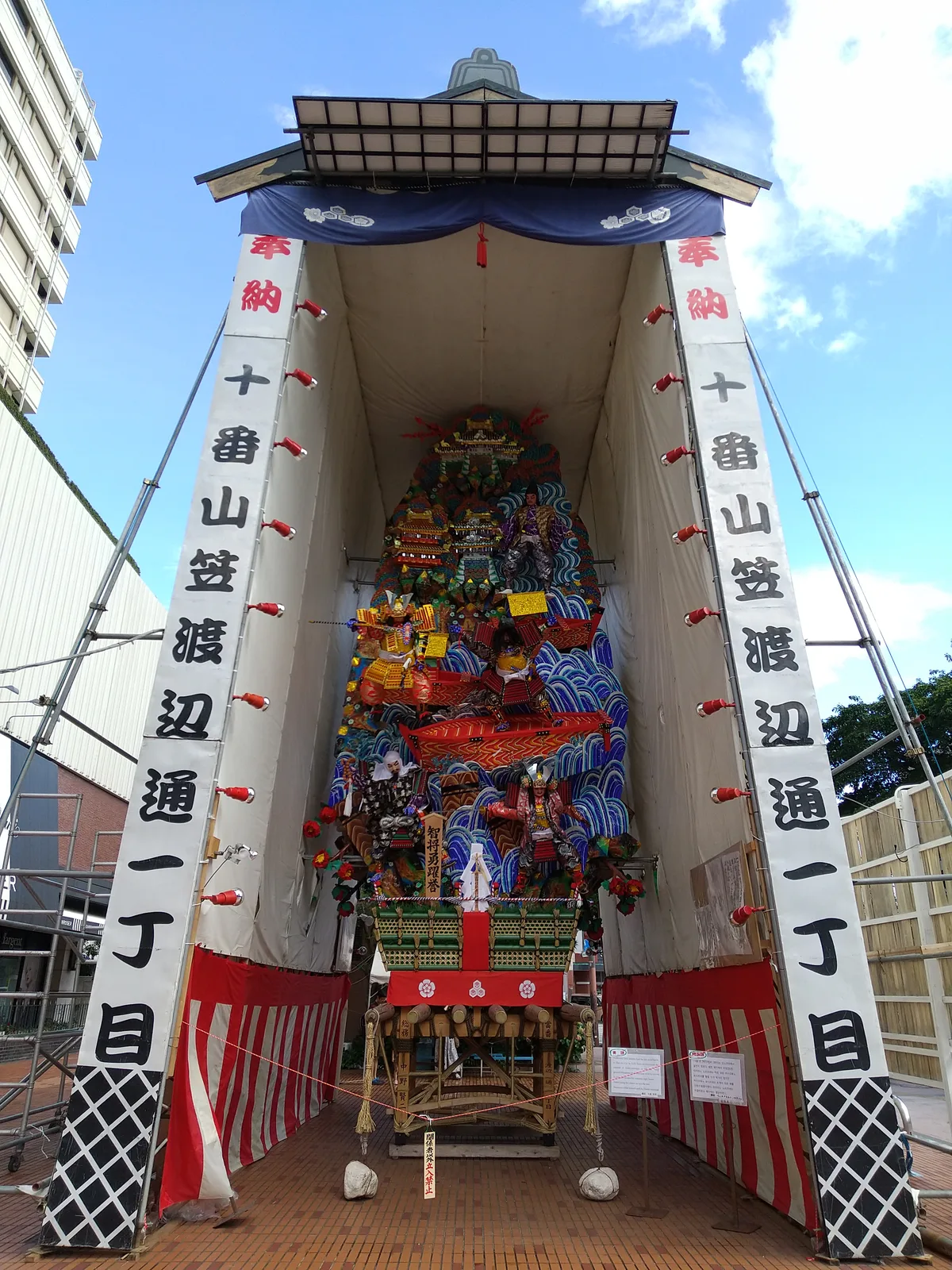 A tall Kazariyama decorated float of Hakata Gion Yamakasa on display in central Fukuoka, covered in samurai figures over a wave motif, photographed in July 2023.