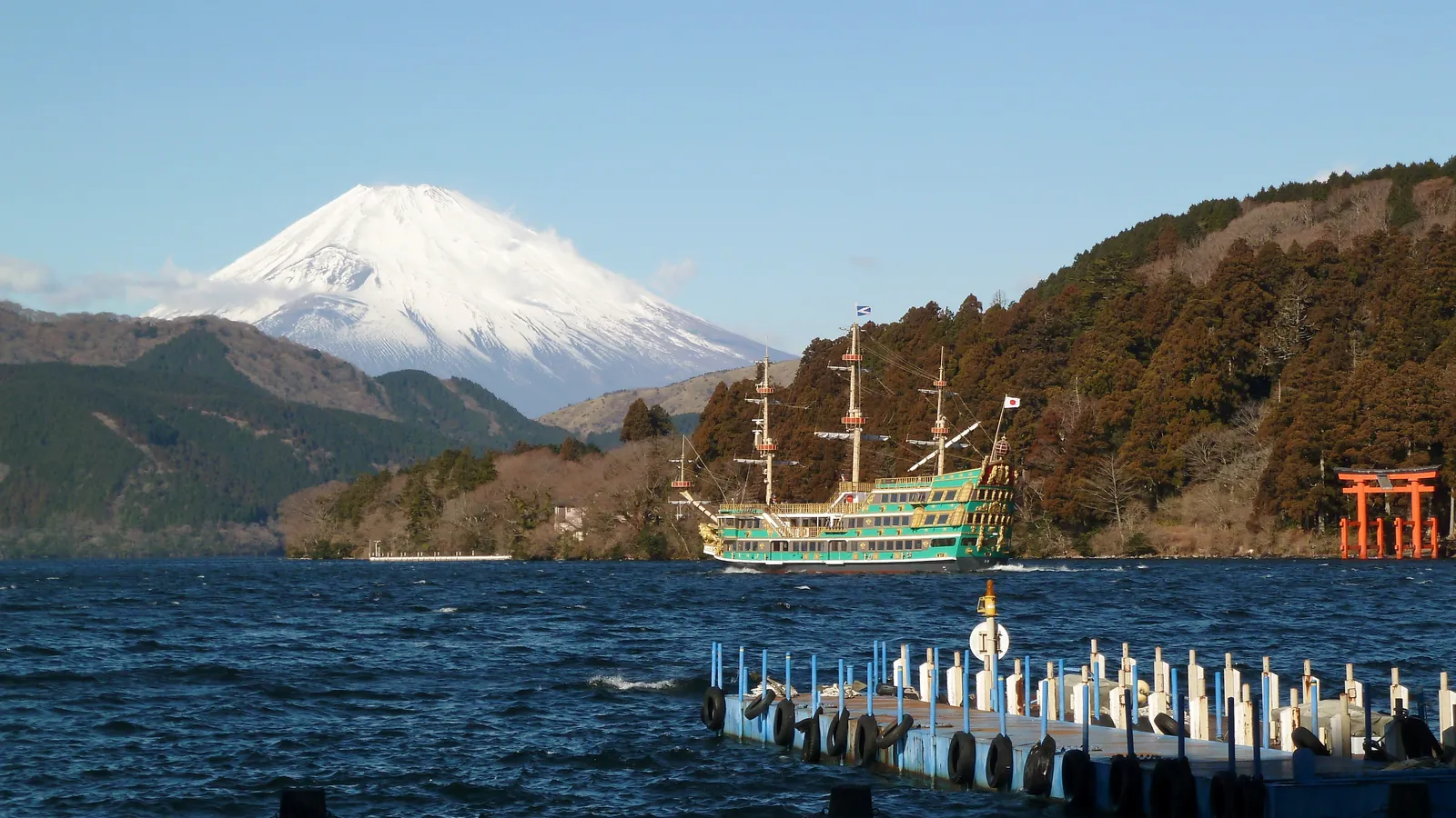 Mt. Fuji seen across Lake Ashi in Hakone, with a sightseeing pirate ship and a red torii gate on the shore.