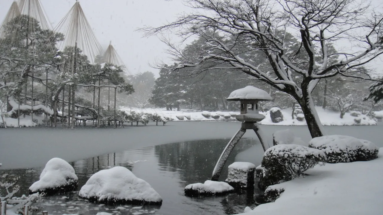 Snow-covered Kenrokuen garden in Kanazawa with a stone lantern on a frozen pond and rope-supported pine trees.