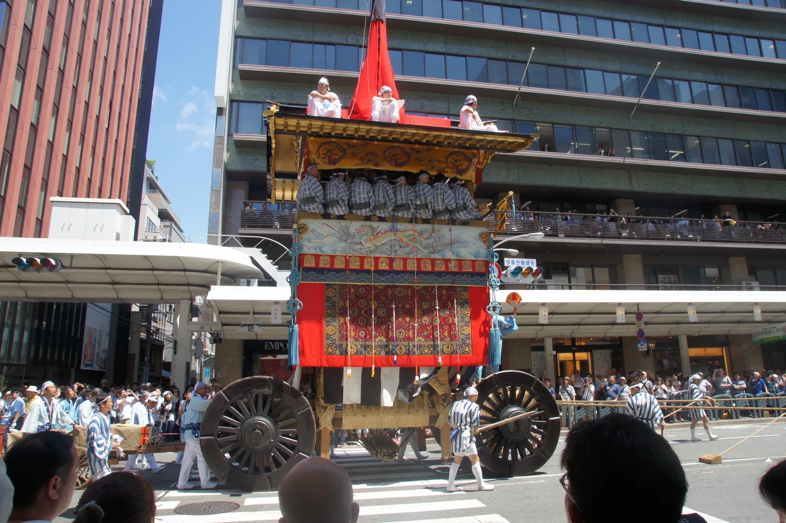 A large yamaboko float with musicians aboard being pulled through a Kyoto street lined with office buildings and spectators during the Gion Matsuri Yamaboko Junko procession.