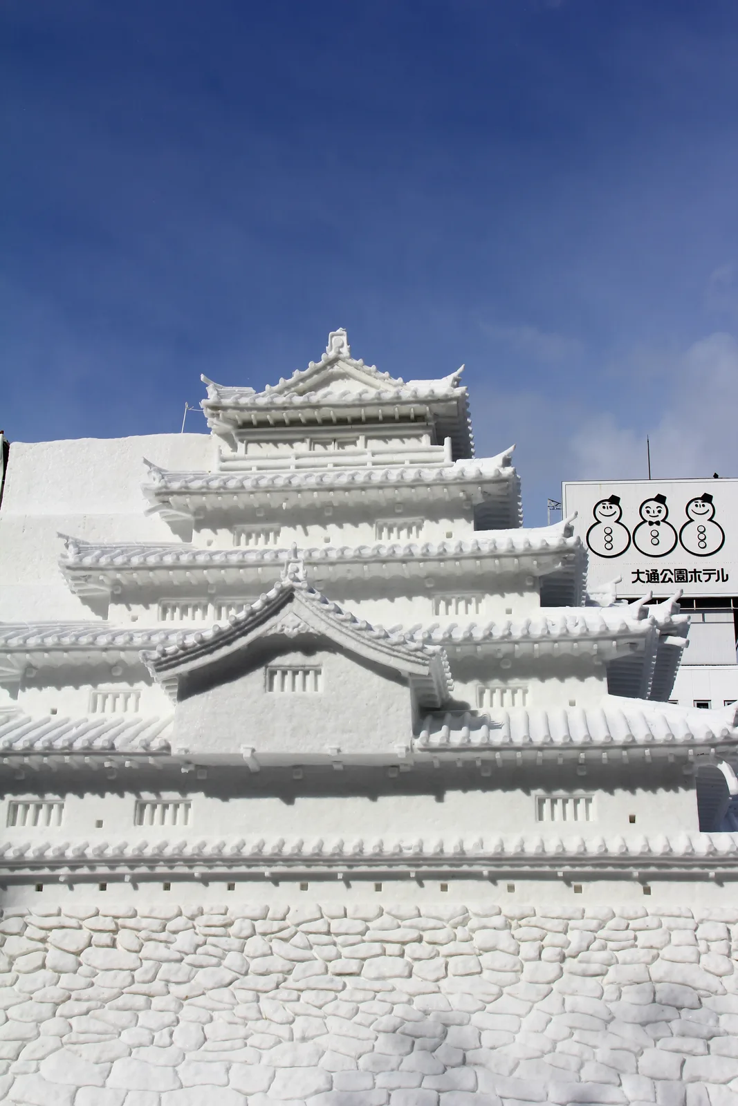 A large snow sculpture shaped like a multi-tiered Japanese castle under a blue sky at the Sapporo Snow Festival's Odori Park site, with a hotel sign visible behind it.