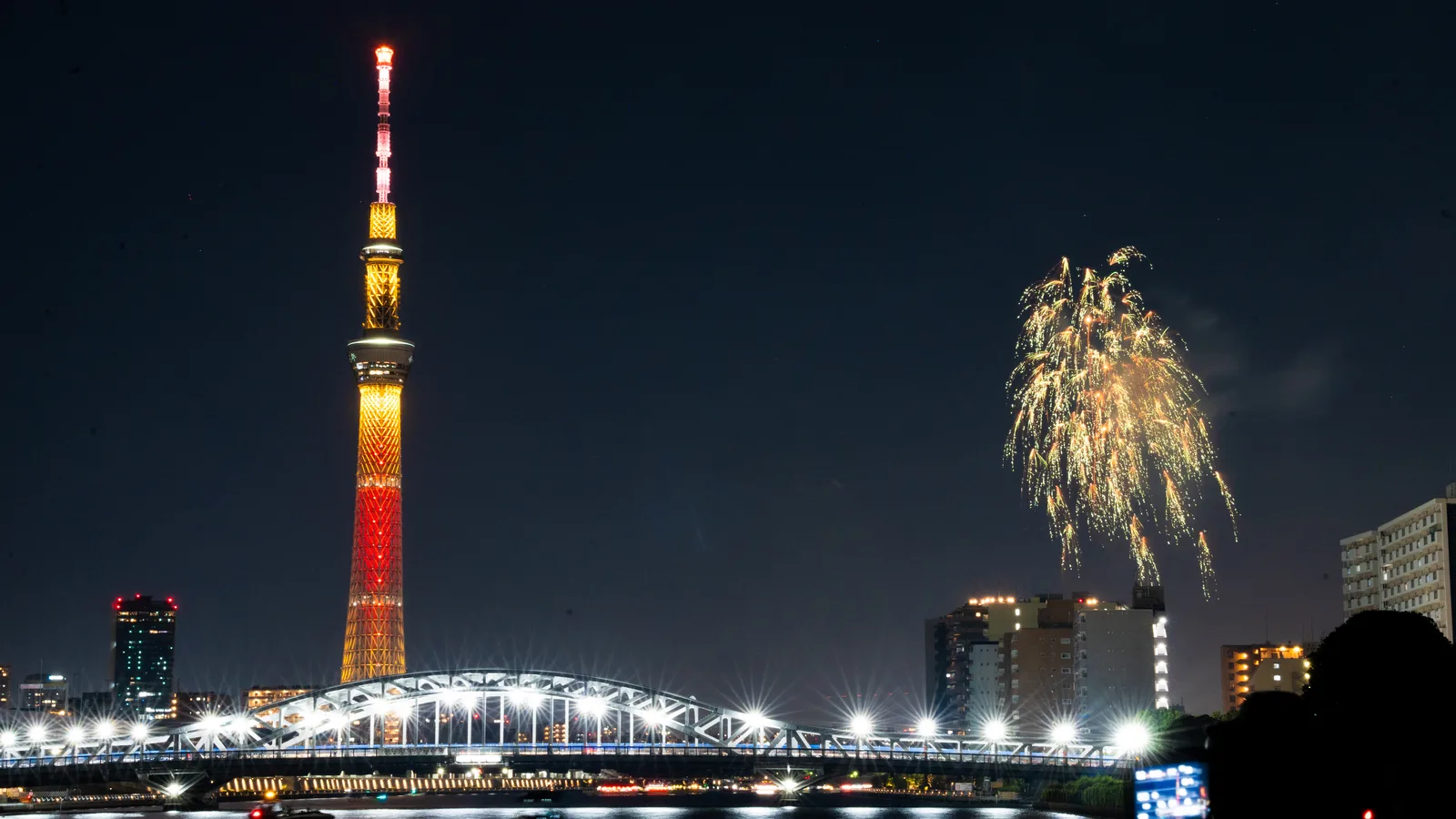 Tokyo Skytree lit in red and gold beside the Sumida River, with a firework burst over apartment buildings during the Sumida River Fireworks Festival, and a lit bridge in the foreground.