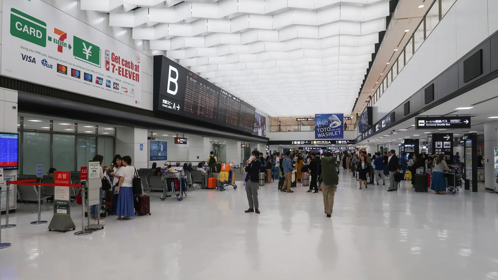 Passengers with luggage carts in the arrival lobby of Narita Airport Terminal 2, beneath a flight information board and a currency exchange sign.