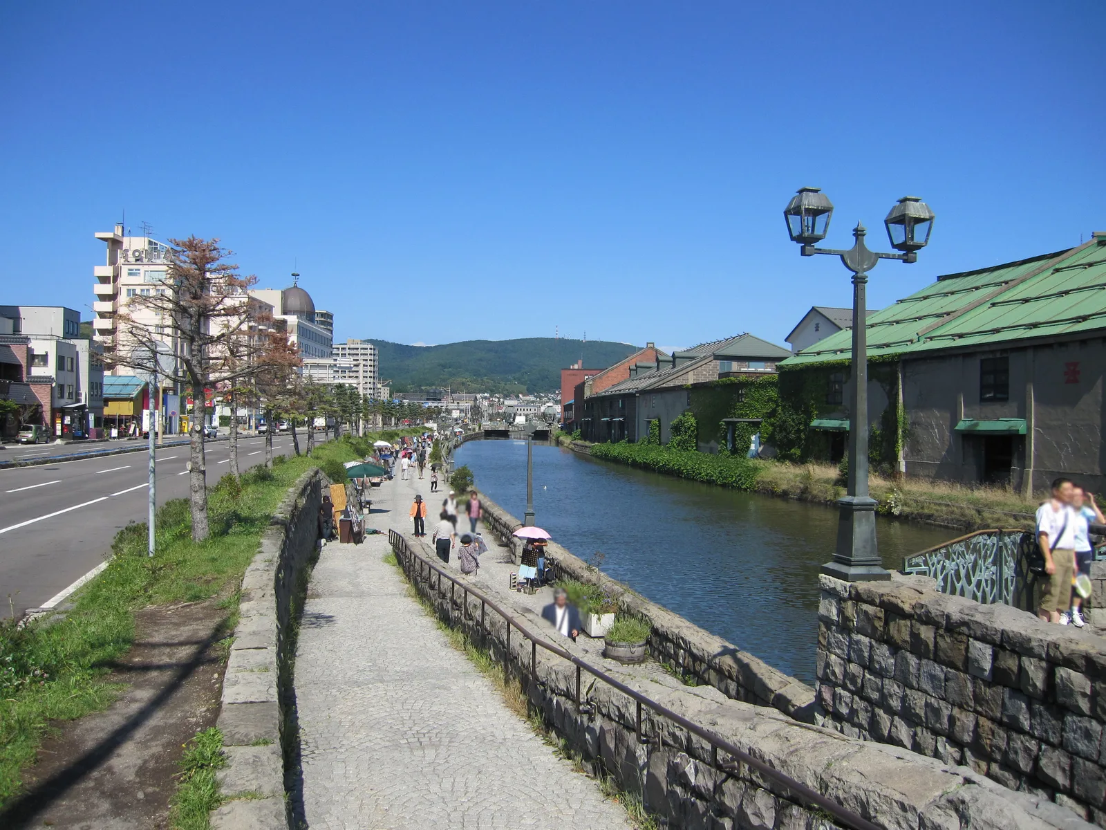 Otaru Canal in summer, lined with old stone warehouses, a stone-paved canal-side promenade and old-style street lamps, under a clear blue sky.