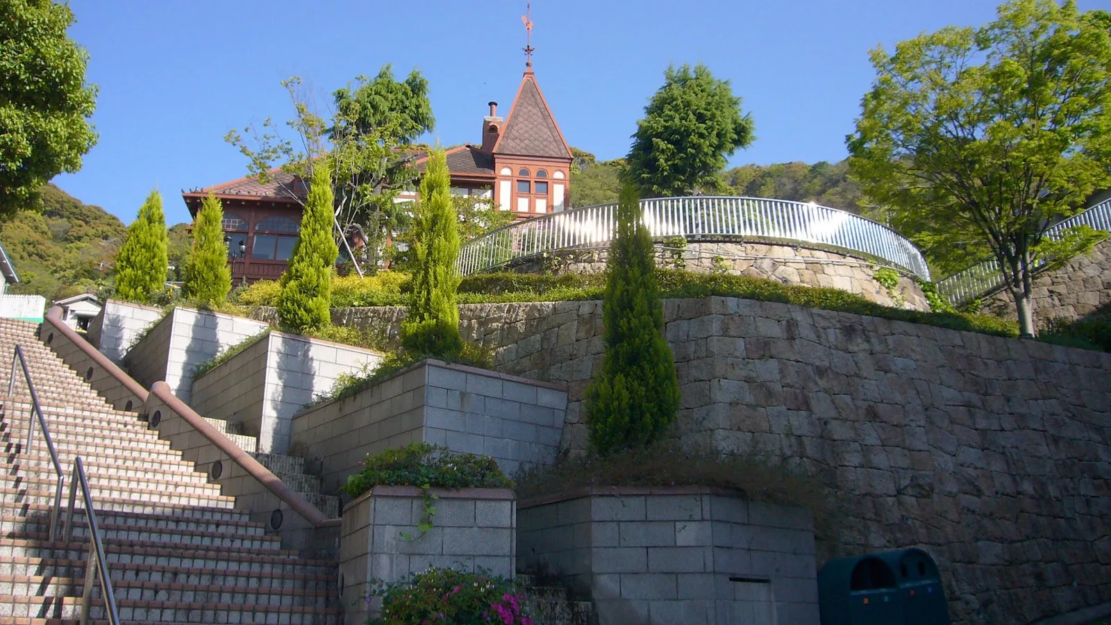 A view from the stone steps below the Weathercock House in Kobe's Kitano-cho, looking up at its red brick tower and weathervane spire framed by trees.