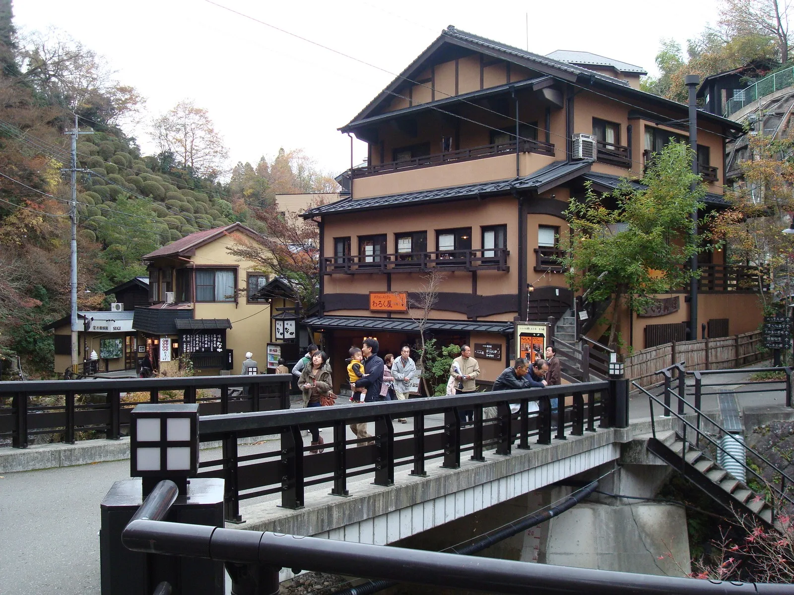 A ryokan and footbridge on a street in Kurokawa Onsen, with a hillside of autumn leaves behind.
