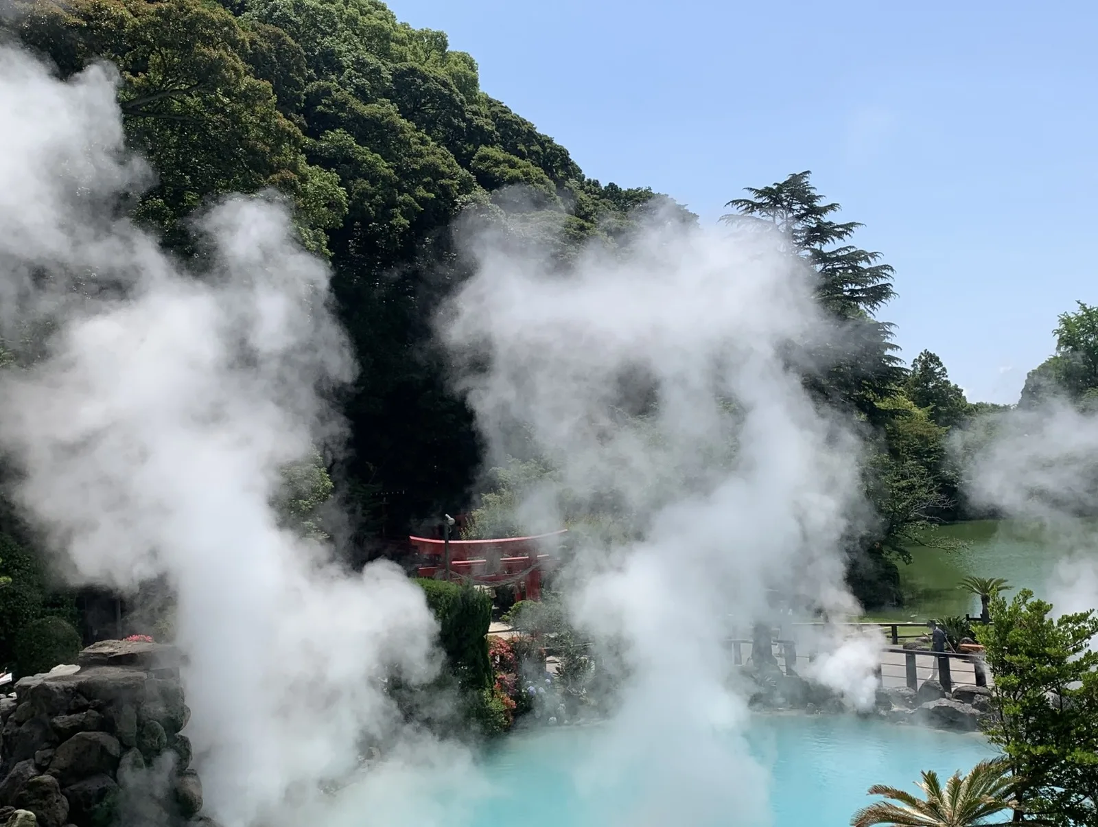 White steam rising from the turquoise hot spring pool at Umi Jigoku in Beppu, with a red torii gate visible through the mist.