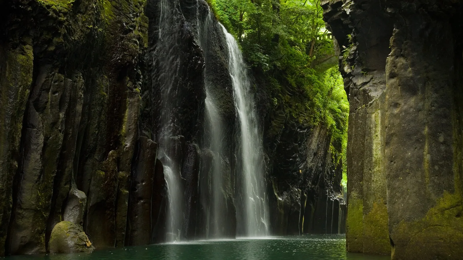 Manai Falls cascading over columnar basalt cliffs into the emerald water of Takachiho Gorge.