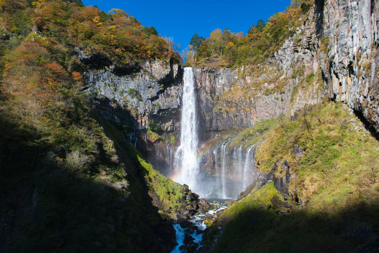 Kegon Falls in Nikko, a tall waterfall dropping into a rocky gorge lined with autumn foliage, with a rainbow visible in the spray below.
