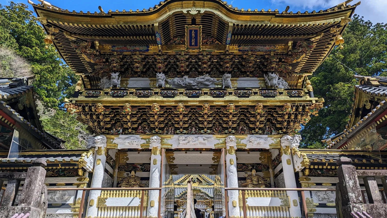 Nikko Toshogu's Yomeimon Gate, a two-story gate covered in gold leaf and carved figures, seen from the stone steps below against a blue sky.