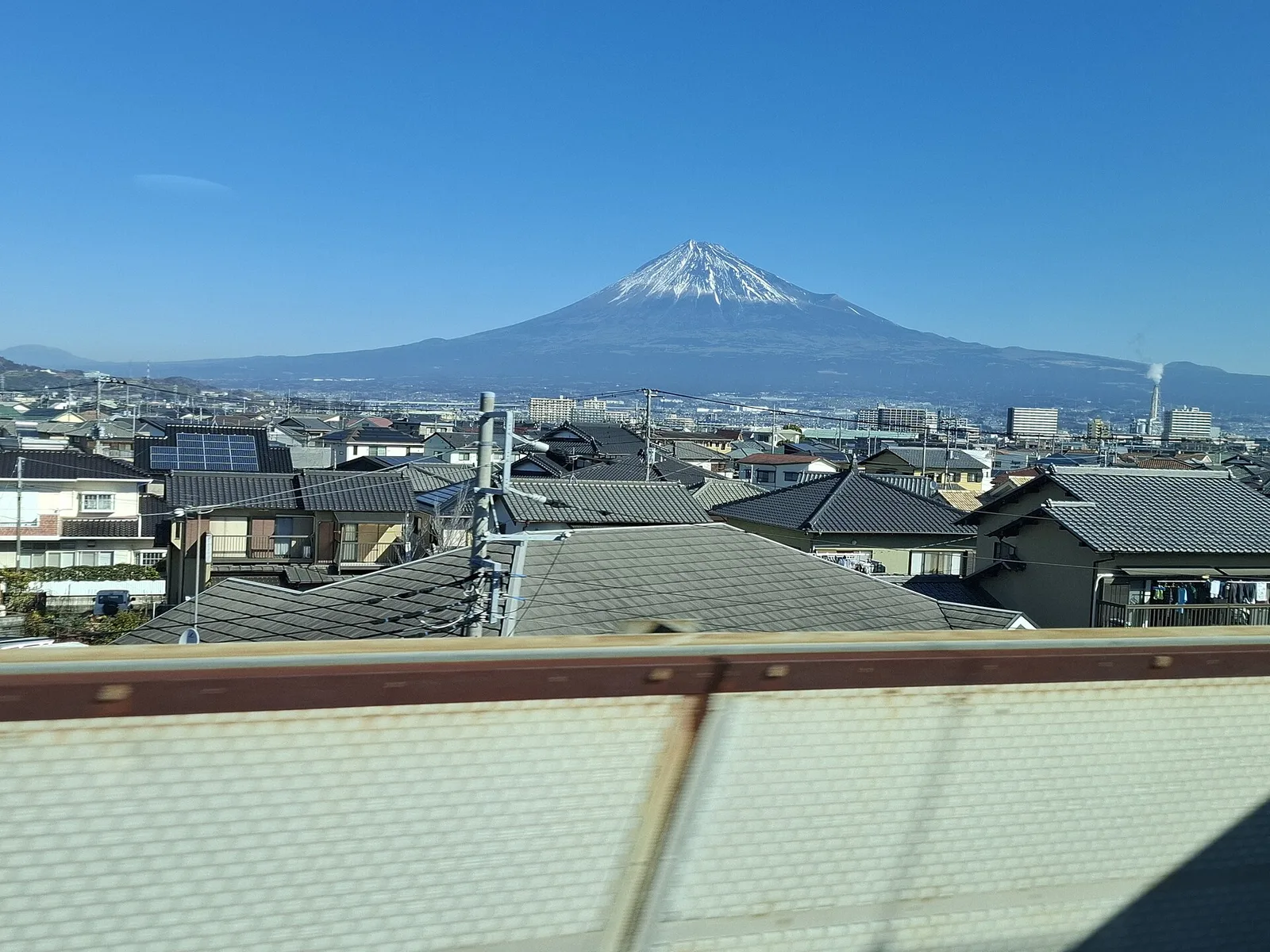 Snow-capped Mount Fuji rising above the rooftops of a town, seen through a train window with the window frame at the bottom.