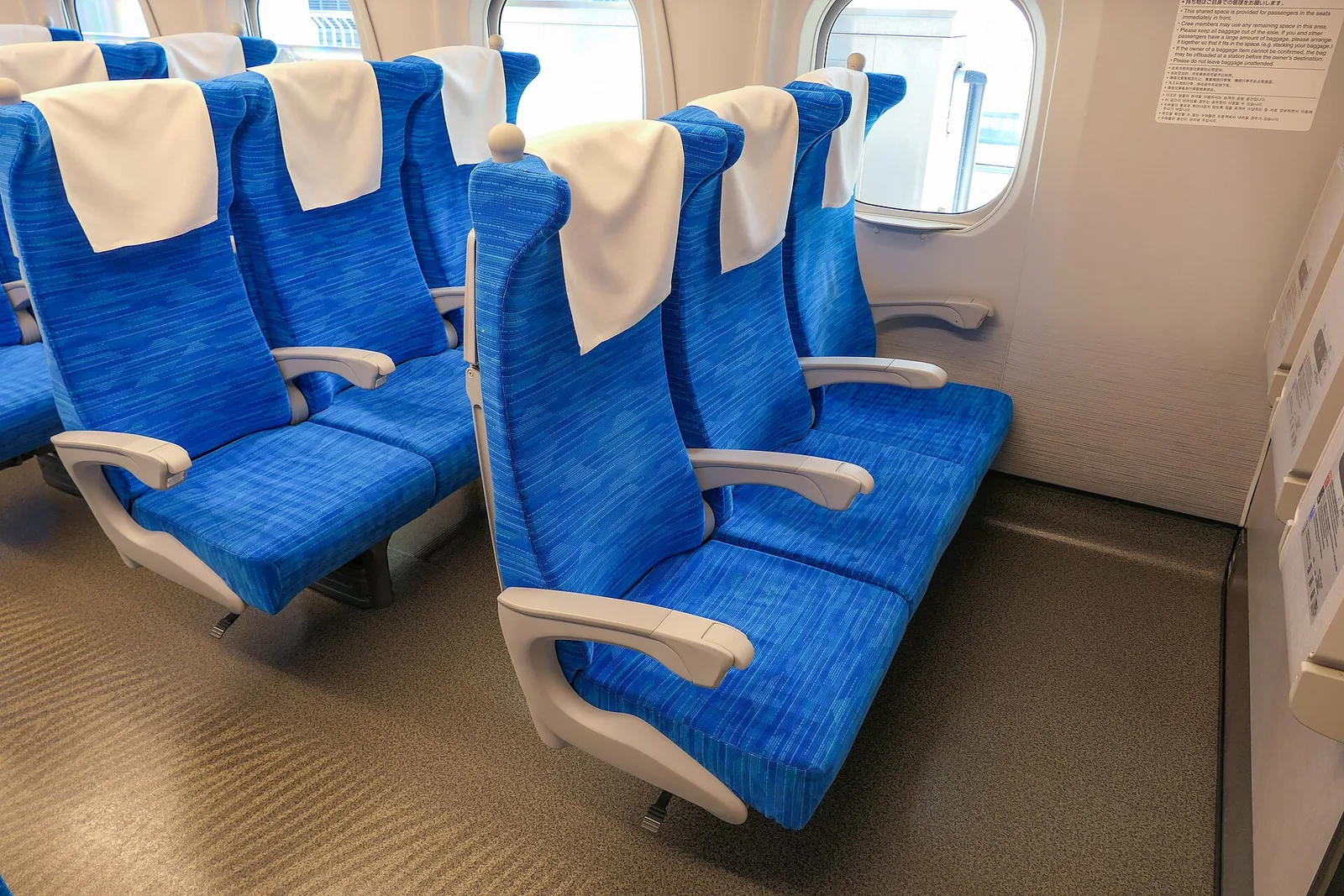 Blue reclining seats in a three-seat row of an N700S ordinary car, with a window and a notice on the wall beside them.