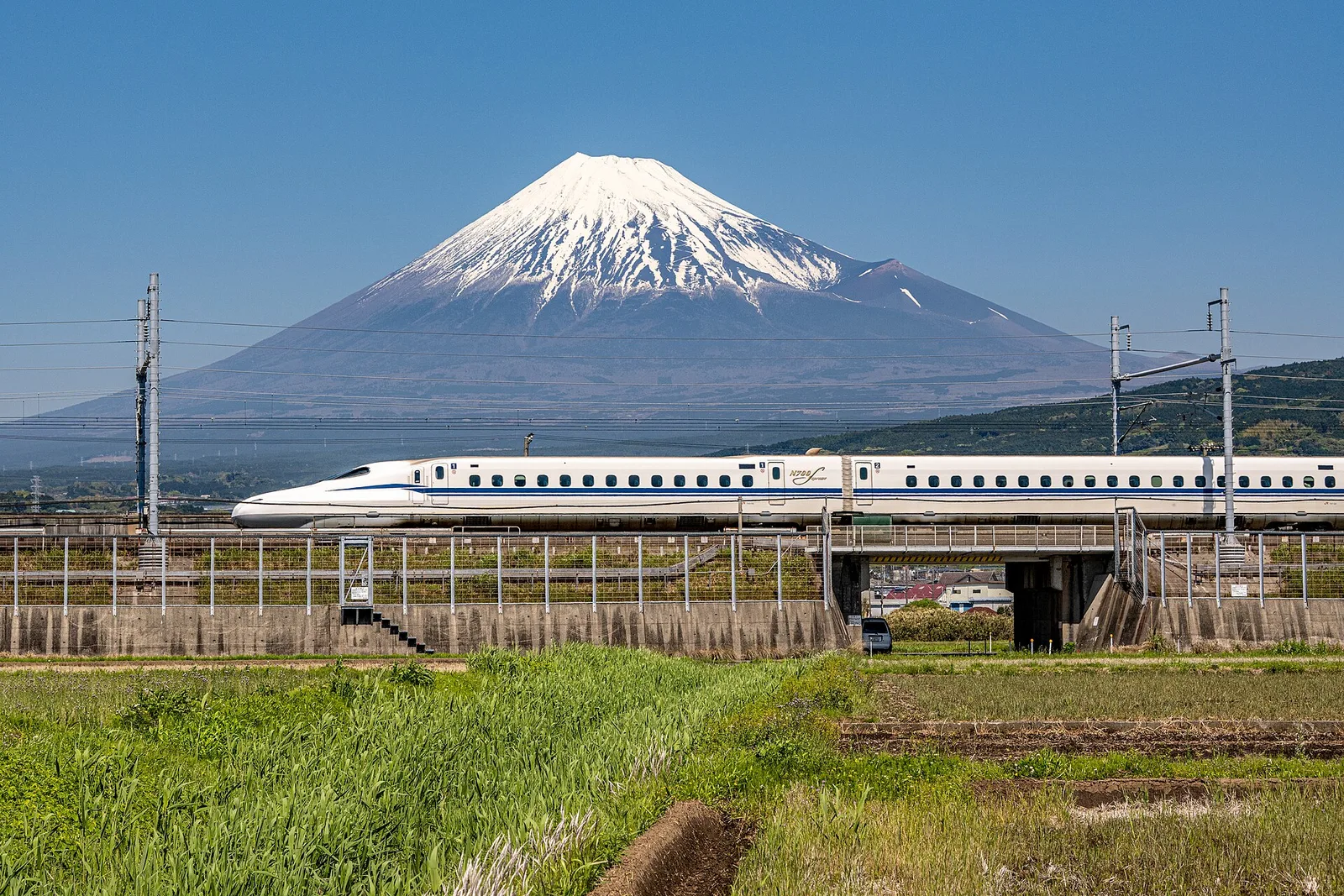 An N700S Tokaido Shinkansen train passing in front of snow-capped Mount Fuji, with fields in the foreground.